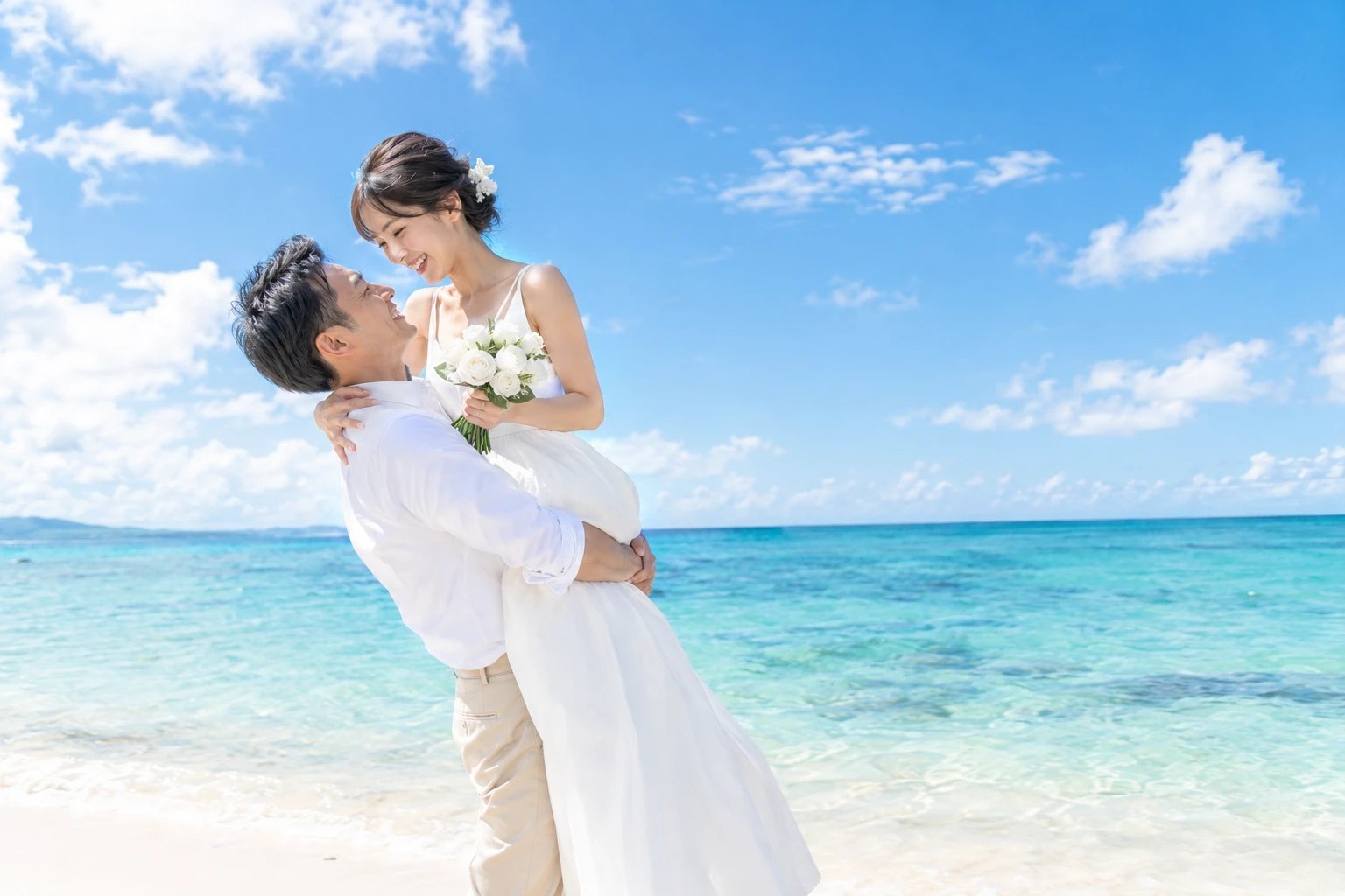 青空の海辺で、白いシャツの新郎がブーケを持った新婦を抱き上げ、二人で笑い合っている